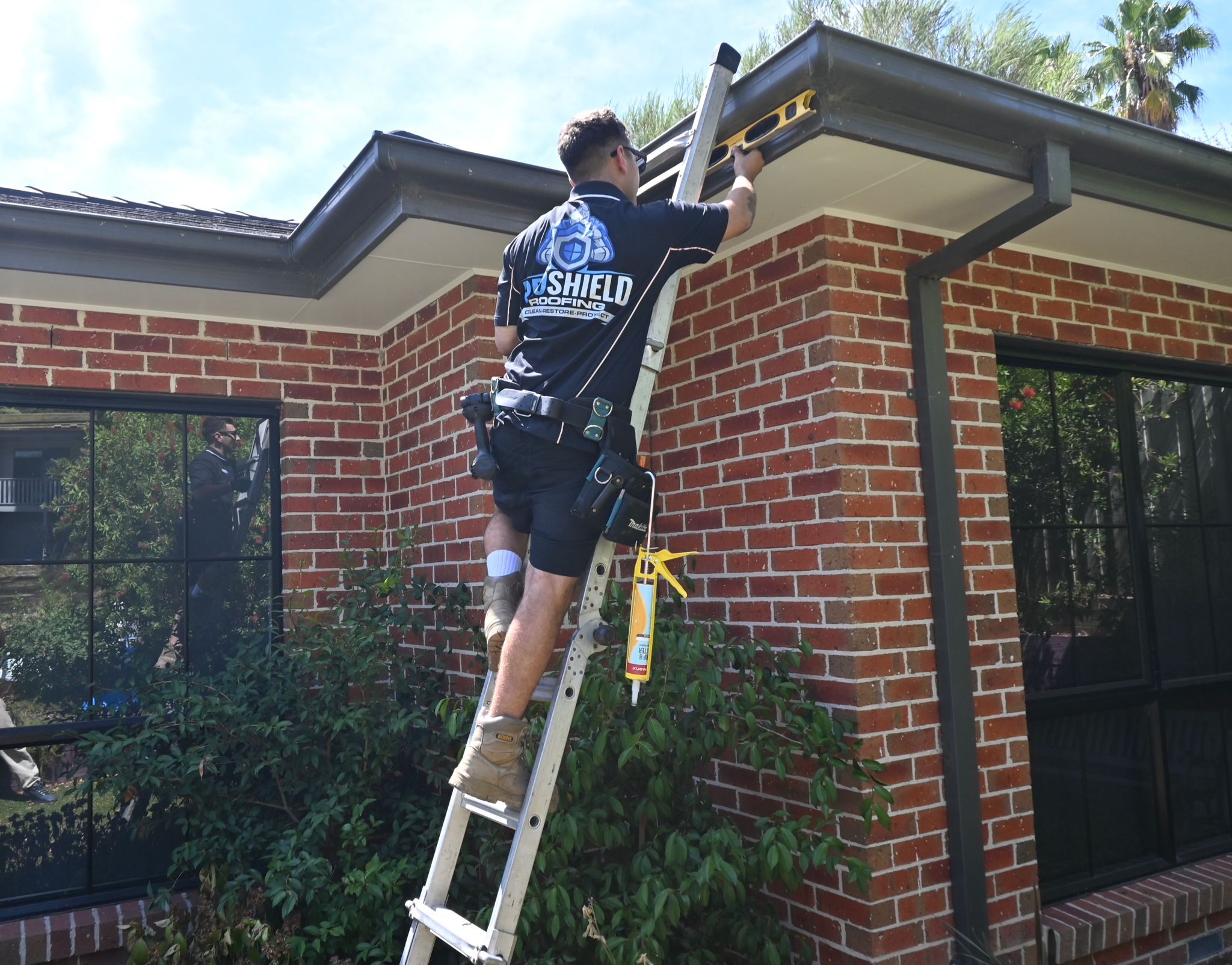 A Pro Shield roofer on a ladder checking a gutter with a level
