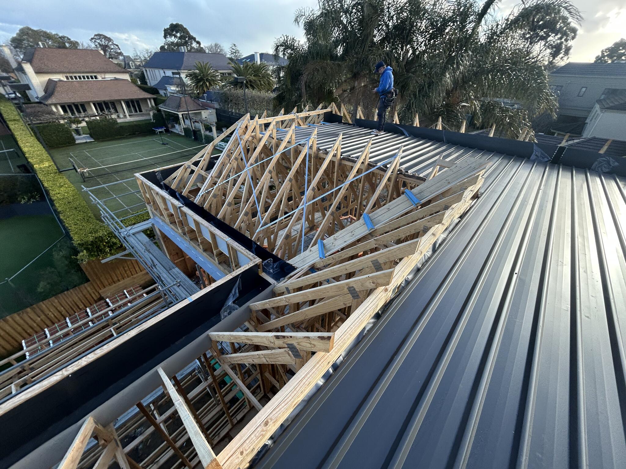 New dark metal roof sheets laid beside open timber trusses, with a roofer at the far edge