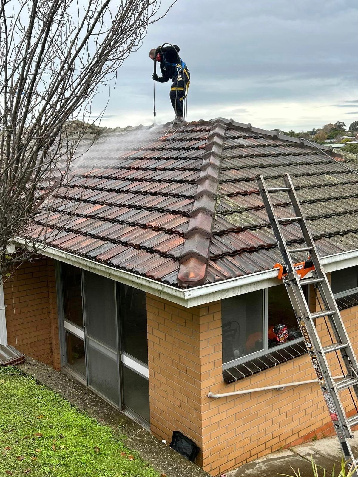 A roofer pressure cleaning a terracotta tile roof on a brick home with a ladder set up