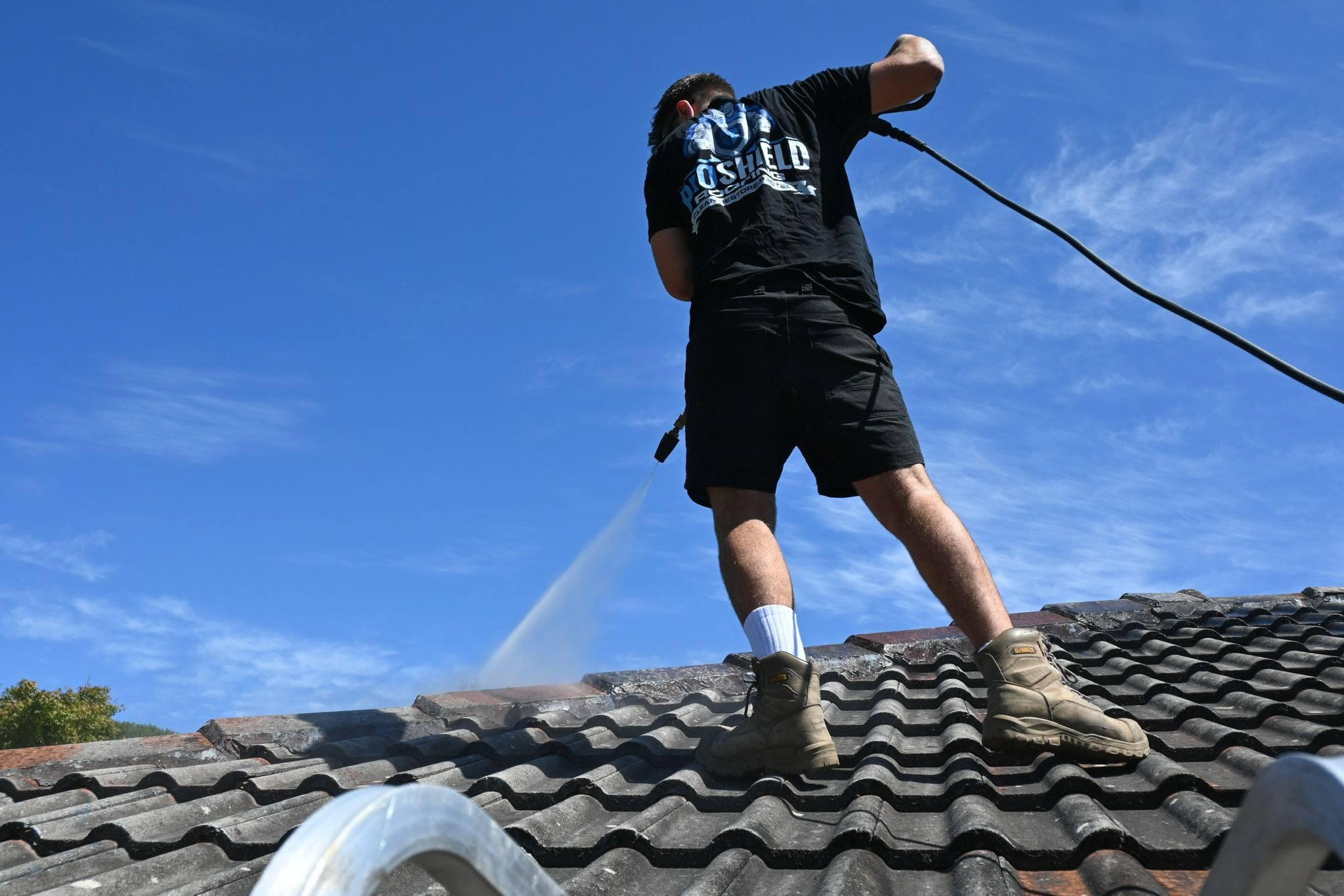 A Pro Shield roofer pressure washing a tile roof under a blue sky